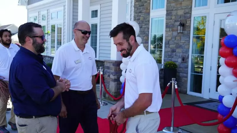 Group of men smiling as one holds pair of large scissors to cut ribbon with